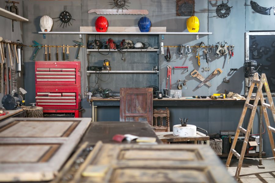 Eine gut sortierte Werkstatt in einer Garage, in der zahlreiche Werkzeuge an der Wand hängen und eine rote Werkzeugkiste im Vordergrund steht. Im Hintergrund befinden sich mehrere Holzarbeiten und eine Leiter, die auf den handwerklichen Charakter des Raum