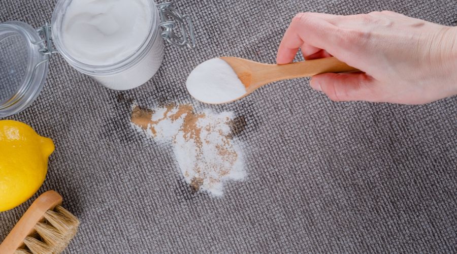 Eine Hand mit einem Holzlöffel streut weißes Natronpulver auf einen braunen Fleck auf einem grauen Teppich. Im Hintergrund sind ein Glasbehälter mit Natron, eine halbe Zitrone und eine kleine Bürste zu sehen. Die Szene illustriert die Verwendung von Hausm