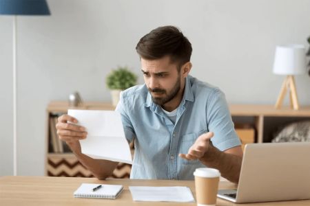Ein Mann mit braunem Haar und Bart sitzt an einem Schreibtisch und hält einen Brief in der Hand – einen Bußgeldbescheid. Er schaut verwundert und ratlos auf das Schreiben. Auf dem Tisch liegen ein Laptop, ein Notizblock mit Stift und ein Pappbecher mit Ka