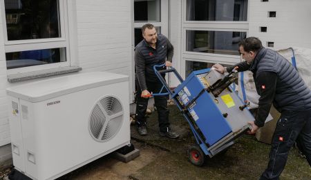 Zwei Handwerker transportieren eine alte Heizanlage auf einer Sackkarre ab, während im Vordergrund bereits das weiße Außengerät einer modernen Wärmepumpe platziert ist.