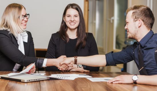 Ein Mann und eine Frau in Business-Kleidung reichen sich lächelnd die Hände über einen hölzernen Konferenztisch. Eine zweite Frau sitzt daneben und beobachtet den Händedruck positiv gestimmt.