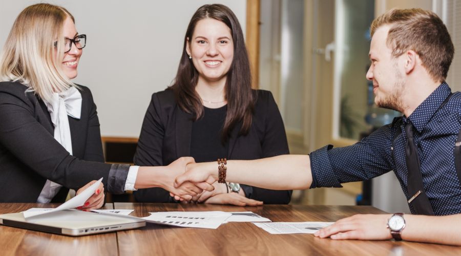 Ein Mann und eine Frau in Business-Kleidung reichen sich lächelnd die Hände über einen hölzernen Konferenztisch. Eine zweite Frau sitzt daneben und beobachtet den Händedruck positiv gestimmt.