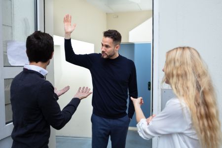 Drei Personen stehen in einer offenen Tür und führen eine hitzige Diskussion mit ausdrucksstarker Gestik. Die Szene wirkt wie eine Konfrontation oder ein lautstarker Streit zwischen Nachbarn.