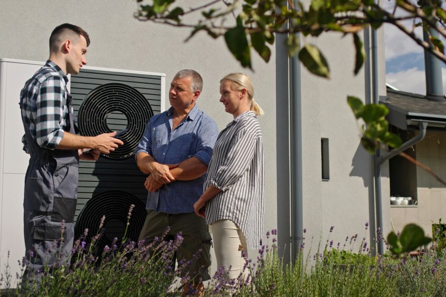 Ein Handwerker in Arbeitskleidung steht im Garten vor einer großformatigen, dunklen Wärmepumpe und berät ein älteres Ehepaar. Die drei Personen besprechen augenscheinlich Details zur Anlage, während der Installateur ein Tablet in der Hand hält.