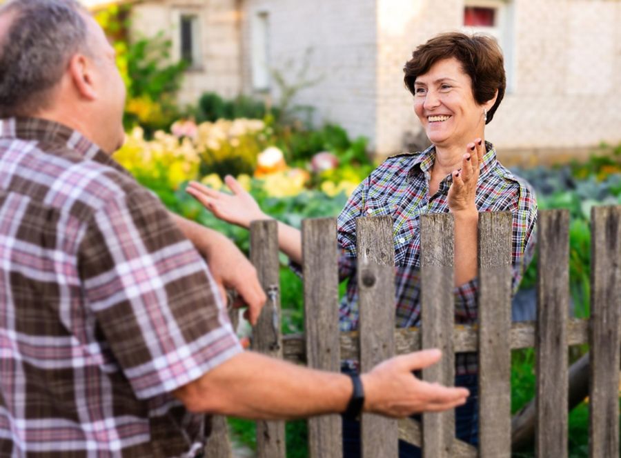 Eine Frau und ein Mann unterhalten sich freundlich über einen hölzernen Gartenzaun hinweg. Beide tragen karierte Hemden. Im Hintergrund ist ein gepflegter Garten mit Blumen und Gemüsebeeten zu sehen.
