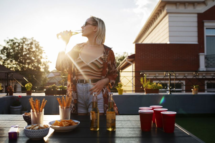Eine junge Frau steht bei Sonnenuntergang auf einer Dachterrasse und trinkt Bier aus einer Flasche. Im Vordergrund steht ein Tisch mit Snacks wie Chips und Knabberstangen sowie roten Bechern.