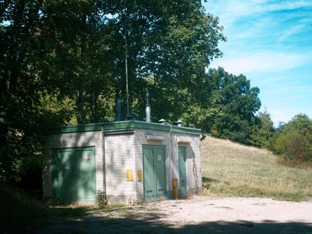 Ein freistehendes, aus hellen Klinkern gemauertes Transformatorenhaus mit grünen Metalltüren und Entlüftungsrohren auf dem Dach steht an einem Schotterplatz. Direkt hinter dem Technikgebäude erstrecken sich dichte Laubbäume und ein sonniger Grashang unter