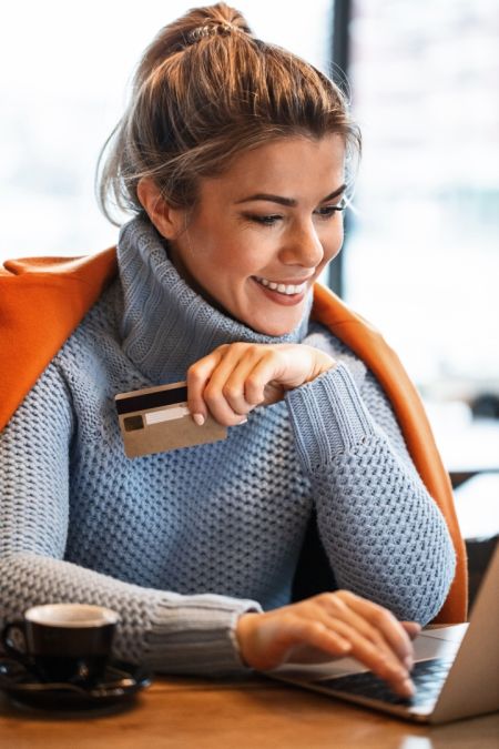 Eine junge Frau sitzt lächelnd an einem Tisch und tippt auf der Tastatur ihres Laptops. In der rechten Hand hält sie eine Kreditkarte bereit, neben dem Rechner steht eine schwarze Kaffeetasse.