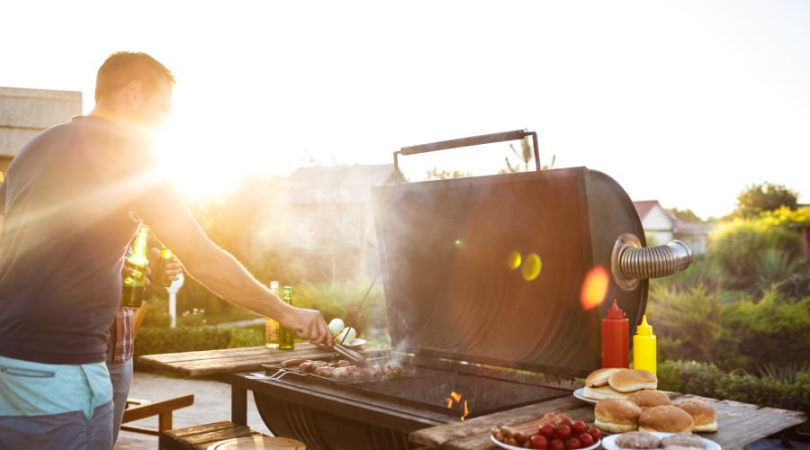 Ein Mann steht an einem großen Barbecue-Grill im Freien, während die warme Abendsonne im Hintergrund scheint. Er wendet Fleisch auf dem Rost mit einer Zange und hält in der anderen Hand eine Bierflasche. Auf einem Holztisch daneben stehen Burgerbrötchen,