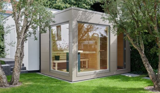 Moderne Gartensauna mit grauer Fassade, großen Panoramafenstern und Flachdach zwischen Bäumen im Garten.