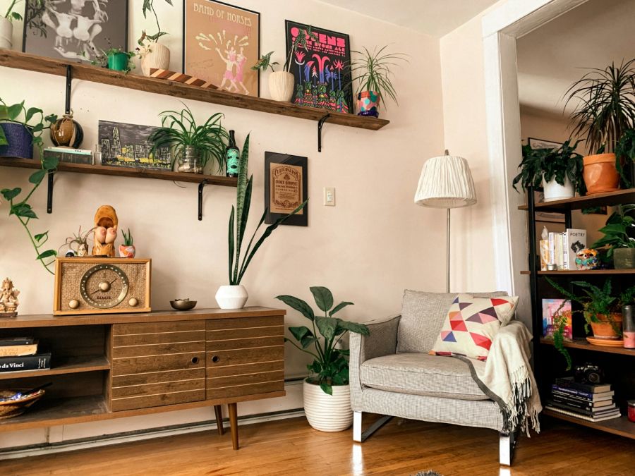 Eine gemütliche Ecke mit einem Mid-Century-Sideboard aus dunklem Holz, auf dem ein altes Röhrenradio steht. Darüber befinden sich Holzregale mit zahlreichen Pflanzen und einer bunten Gallery Wall aus verschiedenen Postern und Kunstdrucken.