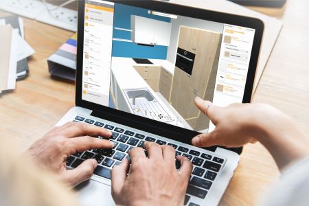 Die Hände einer Person bedienen die Tastatur und das Trackpad eines Laptops. Auf dem Bildschirm ist eine 3D-Planungssoftware für Küchen zu sehen, in der gerade ein Unterschrank mit Backofen konfiguriert wird.