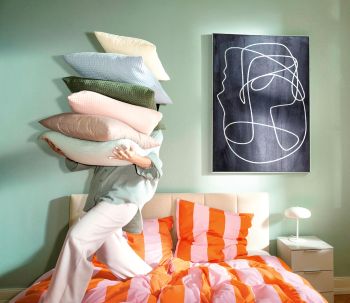 Schlafzimmer mit Bett und Nachttisch, eine Person steht auf Bett und trägt sechs Kissen vor sich als Stapel.