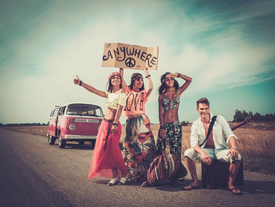 Vier junge Menschen in farbenfroher, gemusterter Kleidung stehen lachend an einer Landstraße neben einem rosa-weißen VW Bulli. Einer hält ein Pappschild mit der Aufschrift „Anywhere“ und einem Peace-Zeichen hoch.