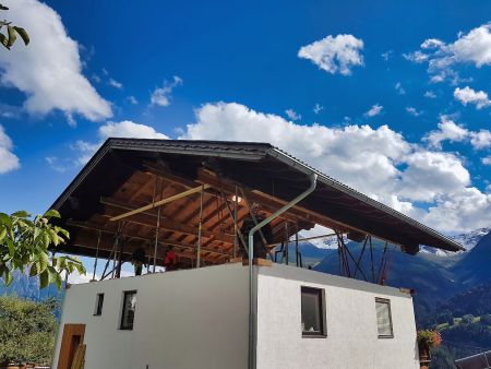 Ein komplettes Satteldach schwebt auf hydraulischen Stützen deutlich über dem gemauerten Erdgeschoss eines weißen Hauses; in der Lücke sind Bauarbeiter und Stützkonstruktionen vor einer alpinen Bergkulisse zu sehen.