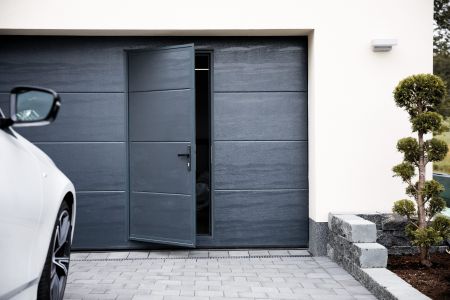 Ein modernes Garagentor, das offen steht, zeigt den Innenraum der Garage. Neben dem Tor steht ein weißes Auto und auf der rechten Seite ist eine kleine, gepflegte Hecke zu sehen.
