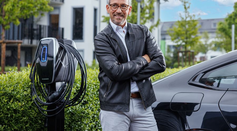 Ein Mann mittleren Alters mit Brille und schwarzer Lederjacke lächelt in die Kamera. Er steht mit verschränkten Armen neben einer modernen Wallbox und einem schwarzen Elektroauto, das in einer gepflegten Wohnsiedlung mit grünen Hecken und modernen Gebäude