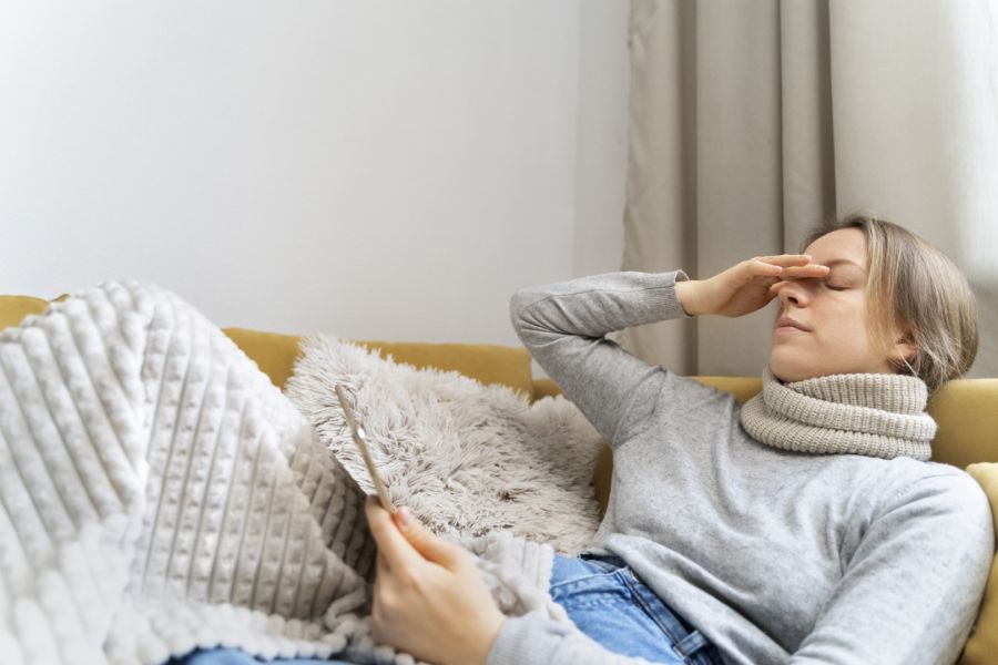 Eine kranke Frau liegt mit einem dicken Rollkragenpullover auf dem Sofa, zugedeckt mit einer Decke. Sie hält sich mit einer Hand die Stirn, was auf Kopfschmerzen oder Fieber hindeutet, während sie in der anderen Hand ein Smartphone hält.