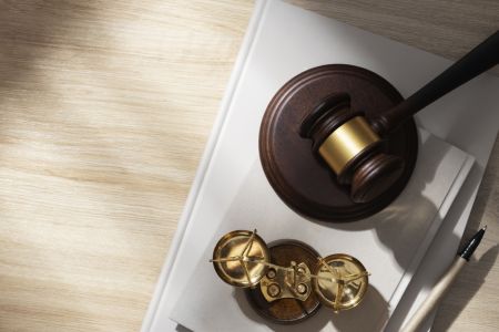 Aus der Vogelperspektive ist ein Holztisch zu sehen, auf dem ein Richterhammer aus Holz, eine goldene Waage der Justitia und ein Stift auf mehreren Büchern liegen. Die Anordnung symbolisiert Rechtsprechung und juristische Urteile.