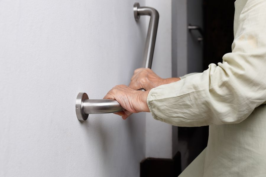 Eine Hand greift nach einem modernen, silbernen Handlauf, der an einer weißen Wand montiert ist. Die Person trägt ein helles, langärmliges Hemd und scheint Unterstützung beim Stehen oder Gehen zu suchen.