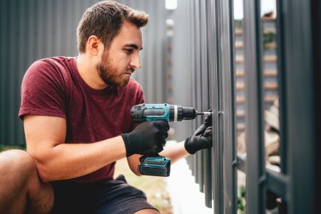 Ein junger Mann mit Bart schraubt mit einem Akkuschrauber eine dunkle Latte an einem modernen Gartenzaun fest. Er trägt schwarze Arbeitshandschuhe und ein bordeauxfarbenes T-Shirt.