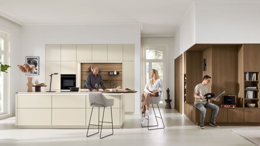 Eine helle, moderne Küche mit einer großen Kochinsel in Beige und Holzoptik. Drei Personen beleben den Raum: Ein Mann steht an der Insel, eine Frau sitzt auf einem hohen Barhocker und ein junger Mann sitzt auf einer integrierten Holzbank.