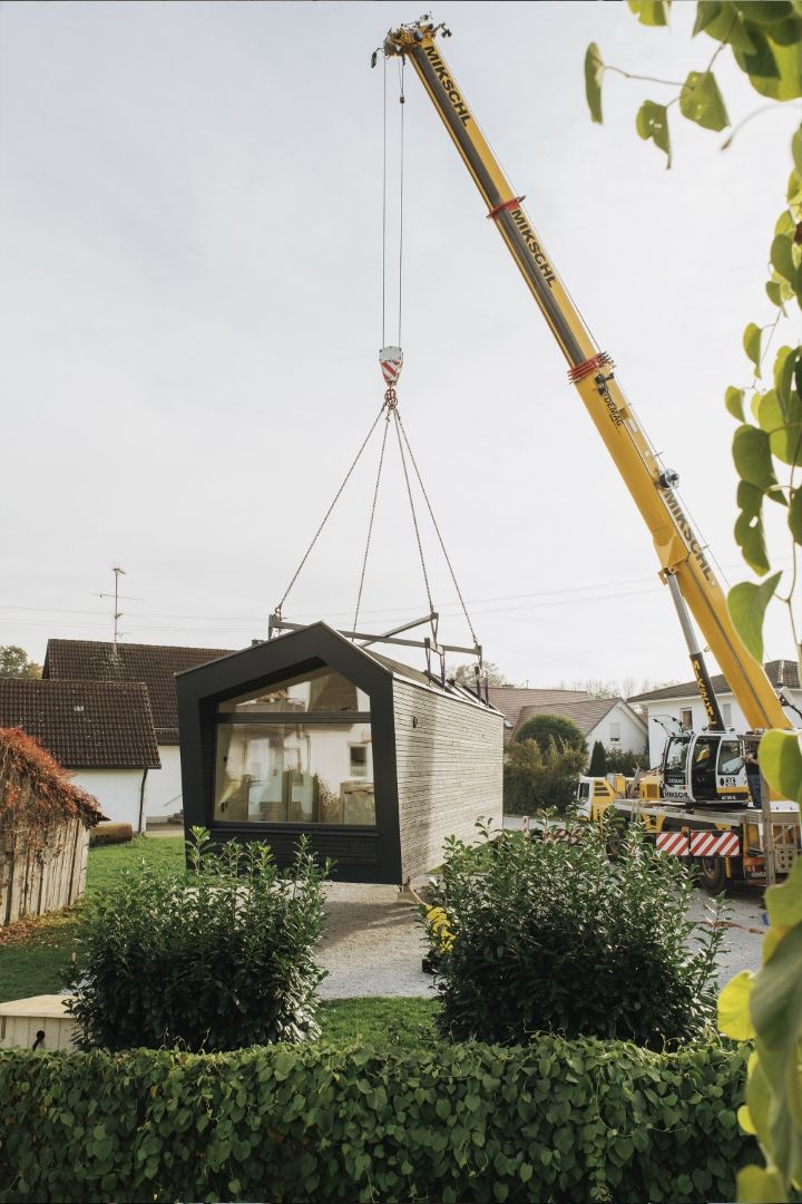 in großes, modernes Tiny-House-Modul wird von einem gelben Kran über ein Grundstück in einem Wohngebiet gehoben. Im Hintergrund sind Dächer anderer Wohnhäuser zu sehen.