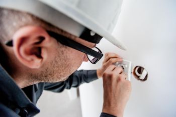 Ein Elektriker mit weißem Schutzhelm und schwarzer Brille arbeitet konzentriert an einer geöffneten Steckdose in einer weißen Wand. Er nutzt seine Hände, um die Kontakte im Inneren der Unterputzdose zu überprüfen oder neu zu installieren.