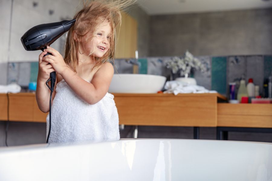 Ein lachendes kleines Mädchen in einem weißen Handtuch steht in einem Badezimmer und föhnt sich die nassen Haare. Sie hält den schwarzen Föhn direkt neben einer weißen Badewanne, was eine potenziell gefährliche Situation darstellt.