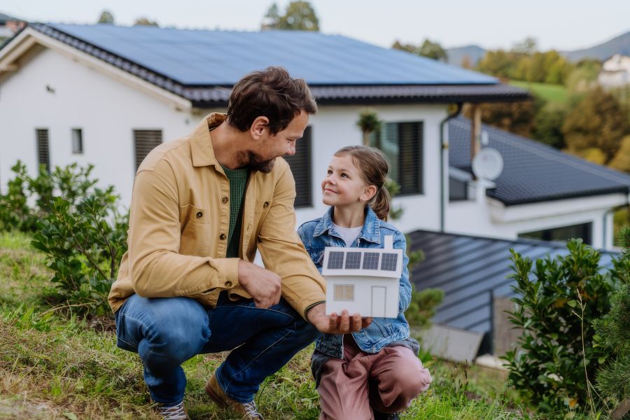 Ein Mann und ein junges Mädchen knien in einem Garten und betrachten gemeinsam ein kleines Modellhaus mit Solarpanels auf dem Dach. Im Hintergrund sieht man ein reales weißes Wohnhaus, dessen Dachfläche fast vollständig mit blauen Solarmodulen belegt ist.