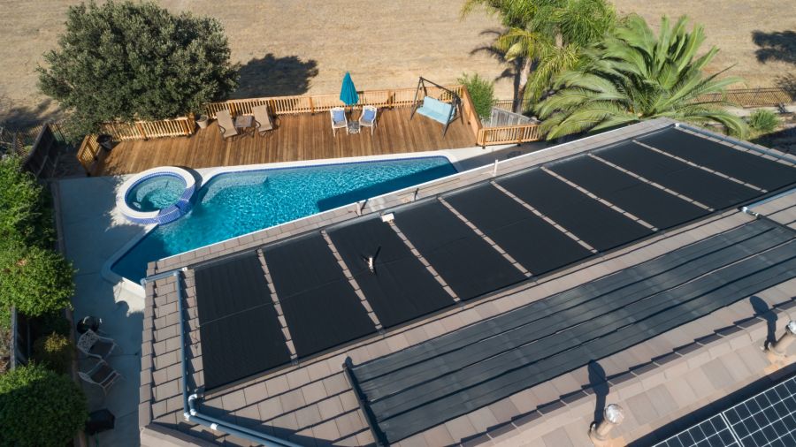 Eine Luftaufnahme zeigt ein Hausdach mit einer großen Solarthermie-Anlage, die direkt über einem nierenförmigen Swimmingpool installiert ist. Neben dem Pool befindet sich eine Holzterrasse, und im Vordergrund ist ein Teil einer Photovoltaik-Anlage zu sehe