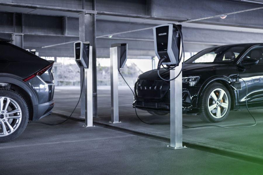 In einer gut beleuchteten Tiefgarage sind mehrere Ladesäulen auf Metallsockeln installiert. Ein schwarzes Elektroauto ist über ein Kabel mit einer der Wallboxen verbunden und lädt, während ein weiteres E-Auto daneben parkt.