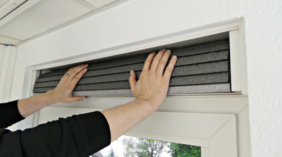 Zwei Hände schieben eine flexible, dunkelgraue und segmentierte Dämmmatte in die Öffnung eines weißen Rollladenkastens. Die Aktion zeigt das Einsetzen der Dämmung über einem Fenster als letzten Schritt der Isolierung.