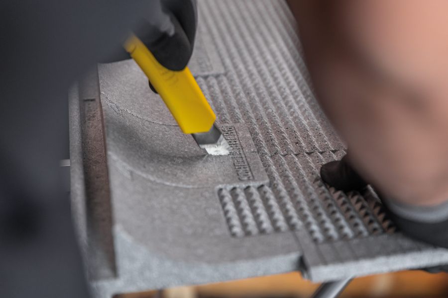 Eine Person mit schwarzen Handschuhen schneidet mit einem gelben Cuttermesser präzise entlang einer vorgegebenen Rille in einem grauen Dämmkeil. Die strukturierte Oberfläche des Materials erleichtert den sauberen Schnitt.