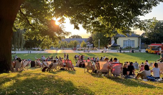 Auf einer Wiese im Abendlicht sitzen viele Menschen gemütlich in Liegestühlen