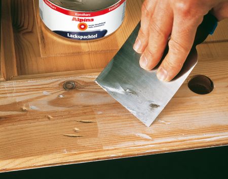 Eine Hand hält einen Metallspachtel und trägt weiße Lackspachtelmasse auf, um tiefe Kratzer in einer hellen Echtholztür zu füllen. Im Hintergrund steht eine geöffnete Dose "Alpina Lackspachtel".