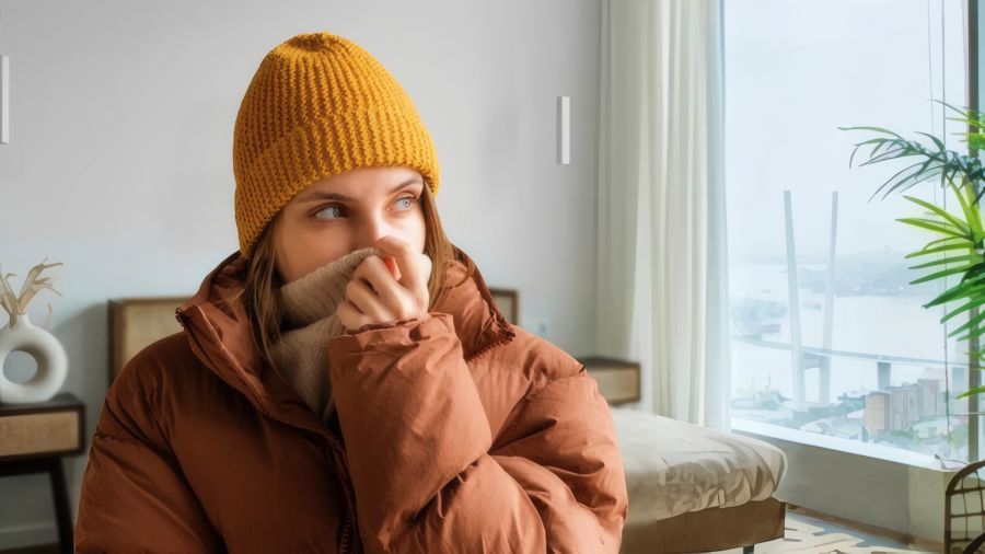 Eine junge Frau mit senfgelber Strickmütze und brauner Daunenjacke zieht fröstelnd den dicken Kragen ihres Pullovers über Mund und Nase. Sie sitzt in einem Wohnraum vor einem großen Fenster mit Ausblick auf eine winterliche Flusslandschaft und eine Brücke