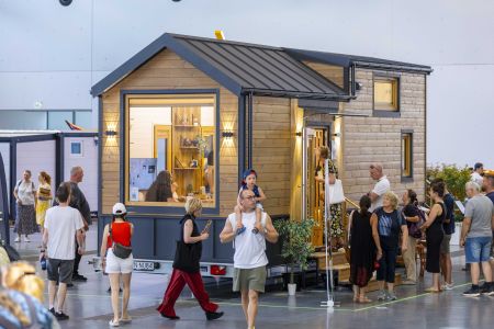 Eine Messehalle, in der ein Tiny House auf Rädern mit Holzfassade ausgestellt ist. Viele Besucher betrachten das Haus, gehen hinein oder stehen davor. Ein Mann trägt ein Kind auf den Schultern.