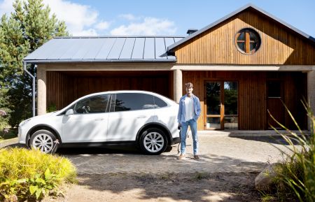 Ein junger Mann steht an einem sonnigen Tag entspannt auf der gepflasterten Einfahrt eines modernen Holzhauses. Er hält das Ladekabel eines weißen Elektroautos, das direkt neben ihm geparkt ist, in der rechten Hand.