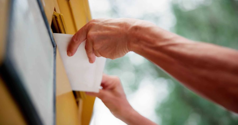 Eine Hand wirft einen weißen Briefumschlag in einen gelben Postbriefkasten im Freien ein.