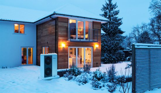 Eine moderne, weiße Luft-Wasser-Wärmepumpe mit zwei übereinanderliegenden Ventilatoren steht aufgestellt im tief verschneiten Garten vor einem Einfamilienhaus. Aus den bodentiefen Fenstern des zweistöckigen Gebäudes mit Holzverkleidung strahlt warmes Lich