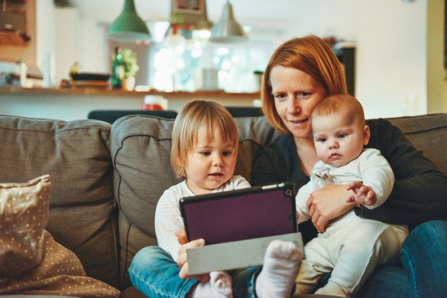 Mutter sitzt mit zwei kleinen Kindern auf einem Sofa und schaut mit ihnen auf ein Tablet.