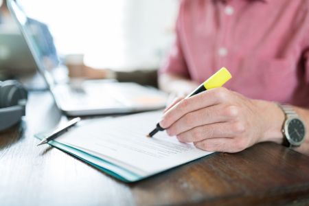 Eine Nahaufnahme zeigt die Hände eines Mannes, der an einem Schreibtisch sitzt und ein gedrucktes Dokument prüft. Mit einem gelben Leuchtstift streicht er konzentriert bestimmte Textstellen auf dem Papier an. Im unscharfen Hintergrund ist ein aufgeklappte