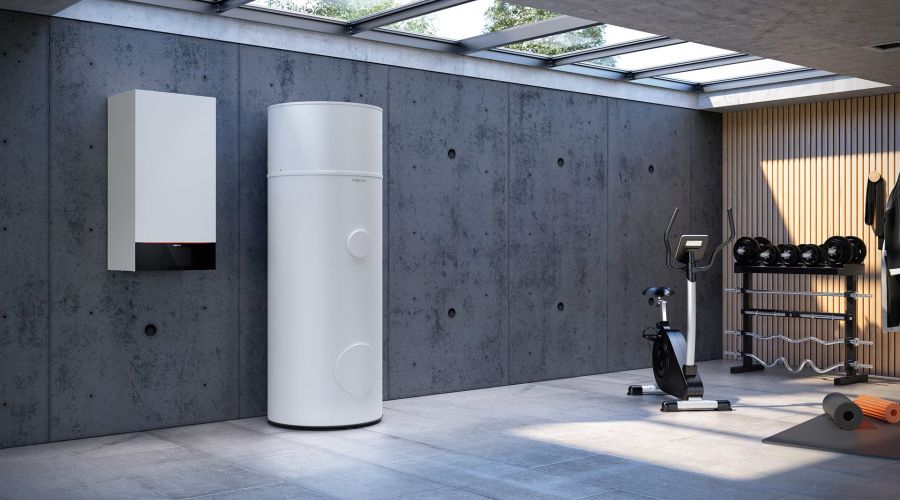 Eine weiße, wandhängende Wärmepumpen-Inneneinheit installiert neben einem hohen, zylindrischen Pufferspeicher in einem modern gestalteten Kellerraum. Der Raum verfügt über Sichtbetonwände, ein Oberlicht und ist teilweise als Fitnessraum mit Hantelbank und