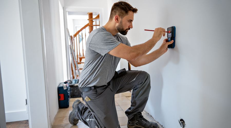 Ein Mann kniet auf dem Boden und markiert mit einem roten Stift durch die zentrale Öffnung eines Ortungsgeräts eine Stelle an der weißen Wand. Das Gerät liegt dabei ruhig an der Wand an, um die exakte Position zu bestimmen.