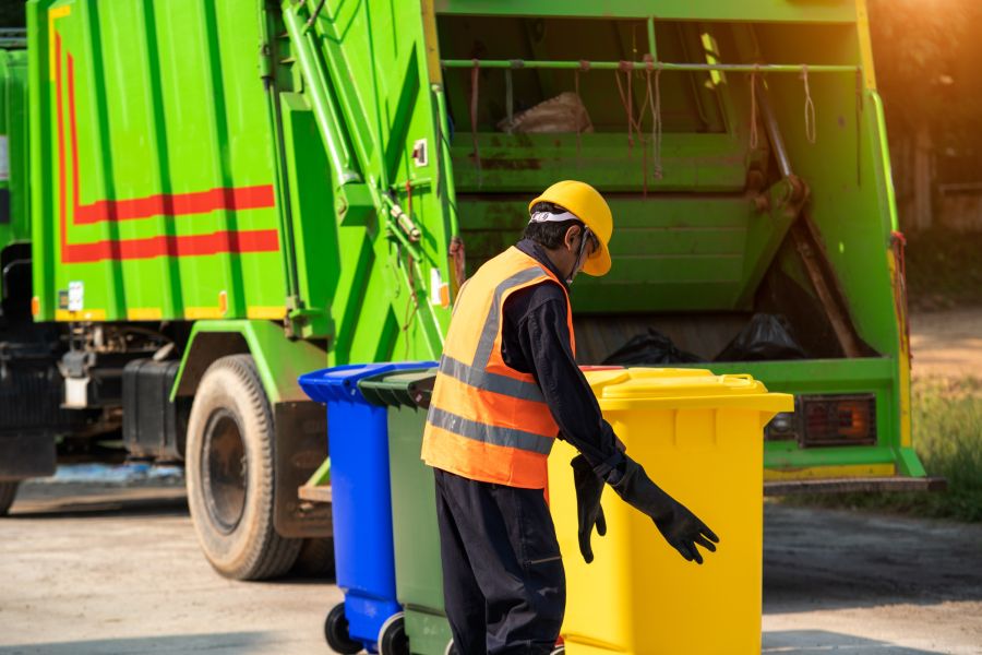 Ein Mitarbeiter in einer orangefarbenen Weste und einem gelben Helm steht neben einem gelben und blauen Müllcontainer, während ein grüner Müllwagen im Hintergrund parkt. Die Szene zeigt eine aktive Abfallentsorgung in einer städtischen Umgebung.