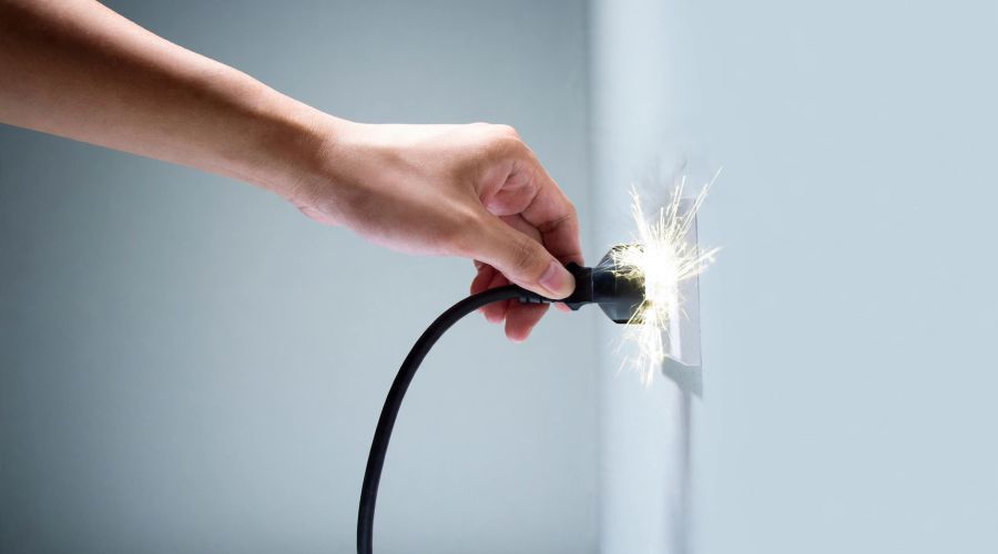 Eine Hand steckt ein schwarzes Stromkabel in eine weiße Wandsteckdose. Dabei entsteht ein heller, sprühender Funkenflug direkt am Kontakt zwischen dem Stecker und der Dose. Die Wand im Hintergrund ist schlicht und in einem hellen Grauton gehalten.