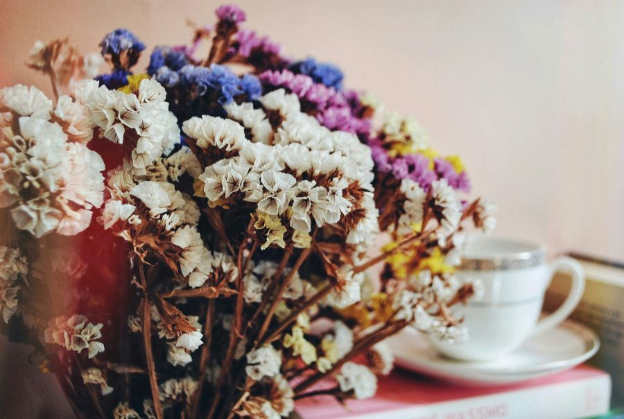 Eine detailreiche Nahaufnahme eines Straußes aus getrockneten Statice-Blüten in Lila und Weiß. Im unscharfen Hintergrund sind eine weiße Kaffeetasse auf einer Untertasse und ein Buch zu erkennen.