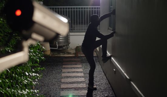 Eine dunkel gekleidete Person mit Gesichtsmaske klettert nachts an einer Hauswand empor, um durch ein Fenster einzusteigen. Im Vordergrund ist eine Überwachungskamera mit einer leuchtend roten Kontrollleuchte unscharf zu sehen.