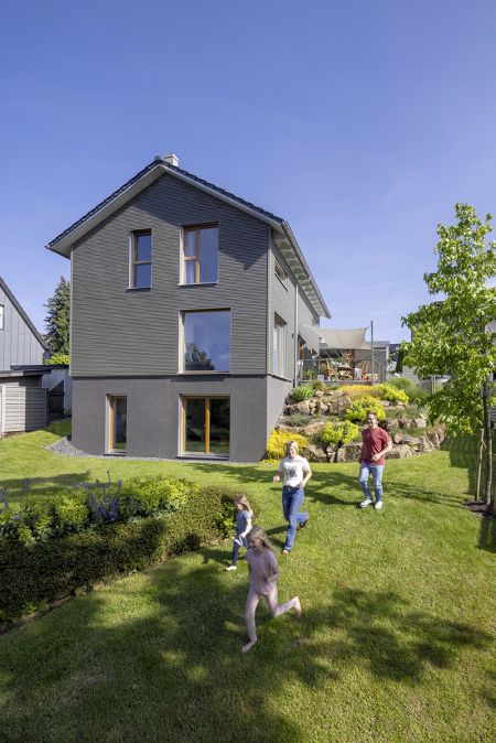 Graues Haus, davor läuft eine junge Familie mit zwei Kindern durch den Garten